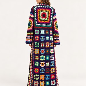 Puede incluir: Un cárdigan largo tipo duster hasta el suelo, confeccionado con un mosaico de cuadrados de ganchillo multicolores. La prenda presenta un color base morado oscuro con acentos vibrantes en rojo, amarillo, verde, azul, naranja y blanco.
