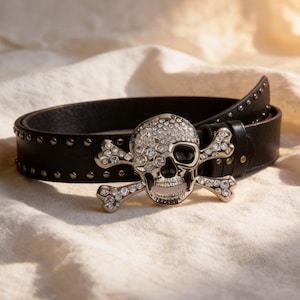 May include: A black leather belt featuring a large silver-toned skull and crossbones buckle encrusted with clear rhinestones. The belt strap is adorned with small metallic studs along the edges, providing a textured appearance to the accessory.