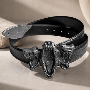 May include: A black belt with a textured surface and a metallic buckle featuring three snarling animal heads. The central head is depicted with an open mouth showing teeth and a tongue, flanked by two side heads with bared fangs. The belt strap has a crocodile-like embossed pattern and a pointed metal tip.