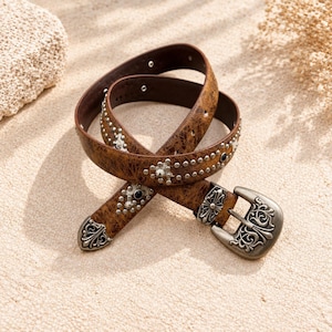 May include: A brown distressed leather belt featuring silver-toned metal studs, decorative floral-patterned metal accents, and a matching ornate silver-toned buckle. The belt strap has a weathered texture and includes small circular metal rivets.