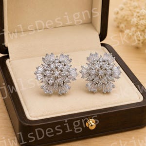 May include: A pair of silver-toned stud earrings featuring a cluster of clear, faceted stones arranged in a floral or snowflake pattern. The earrings are displayed inside a dark brown wooden box with a cream-colored velvet interior lining.