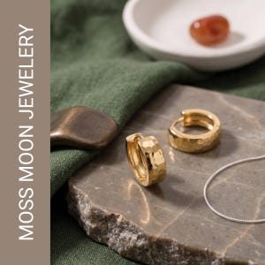 Puede incluir: Un par de pendientes de aro dorados con una superficie martillada y texturizada. Los pendientes se muestran junto a una cadena fina de color plateado. El texto MOSS MOON JEWELERY es visible en el lado izquierdo de la imagen.