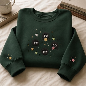 May include: A dark green crewneck sweatshirt featuring embroidered black soot sprites with large white eyes. The design includes small colourful stars in yellow, pink, green, and white scattered around the sprites on the chest and on each sleeve.