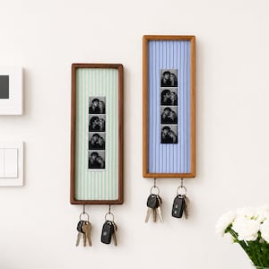 May include: Two rectangular wooden wall-mounted key holders with fabric backings. The left holder has mint green striped fabric with a black and white photo strip. The right holder has blue striped fabric, a photo strip, and two metal key hooks.