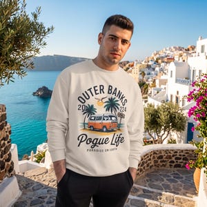 May include: A cream-colored long-sleeve sweatshirt featuring a graphic print of an orange and white vintage van, palm trees, a sun, and the text Outer Banks 2024 Pogue Life Paradise on Earth. The garment has a crew neckline and ribbed cuffs.