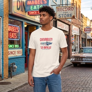 Op de afbeelding: Een crèmekleurig T-shirt met korte mouwen en een afbeelding van een blauwe Chevrolet Impala uit 1959. De tekst luidt CHEVROLET! '59 IMPALA! ALL NEW ALL OVER AGAIN! Het T-shirt heeft een ronde hals en is gemaakt van een effen, lichtgekleurde stof.
