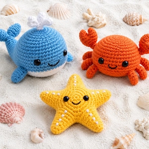 May include: A blue crocheted whale with a white spout, an orange crocheted crab with two claws, and a yellow crocheted starfish with white accents. Each plush toy features black safety eyes and a stitched smile, arranged on a sandy surface.