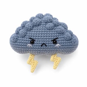 Op de afbeelding: Een blauwe gehaakte wolk als knuffel met een textuurrijk, bobbelig oppervlak. Het heeft een norse gezichtsuitdrukking met zwarte kraaloogjes, een klein fronsend mondje en witte stiksels. Twee gele gehaakte bliksemschichten hangen onderaan.