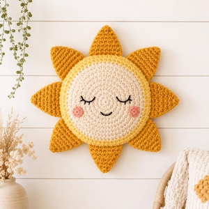 以下が含まれることがあります： クリーム色の中心部に刺繍された顔、閉じた目、ピンクの頬を持つ、かぎ針編みの太陽の形をした壁掛け装飾。太陽には8つの尖ったマスタードイエローの光線が外側に放射状に伸びており、すべて質感のある毛糸で丁寧に作られています。