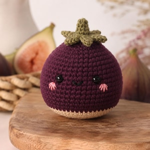 Puede incluir: Un higo amigurumi de ganchillo, redondo y de color morado oscuro, con una base de color beige claro. Presenta un tallo verde de ganchillo en la parte superior, dos pequeños ojos de cuentas negras, una boca negra bordada y mejillas rosadas.