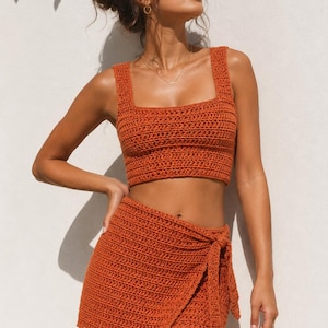 May include: A burnt orange two-piece crochet set consisting of a sleeveless crop top with a square neckline and a matching wrap-style mini skirt featuring a side tie closure. The garment is crafted with a consistent horizontal stitch pattern.