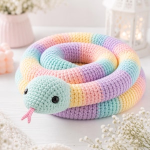 May include: A coiled crochet snake toy featuring a pastel rainbow color palette of light blue, yellow, pink, and lavender yarn. The snake has two black plastic eyes and a small, forked pink tongue protruding from its rounded head.