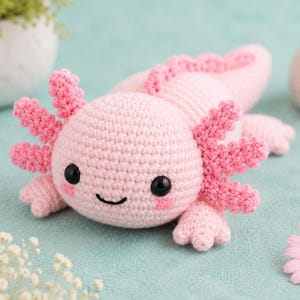 Könnte beinhalten: Ein hellrosa gehäkeltes Axolotl-Plüschtier mit rundem Kopf, schwarzen Kunststoffaugen und einem kleinen gestickten Mund. Es verfügt über drei verzweigte äußere Kiemenpaare an jeder Kopfseite, einen langen Schwanz und vier kleine Gliedmaßen mit Zehen.