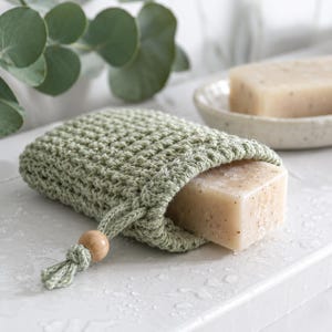 May include: A sage green crocheted soap pouch with a drawstring closure featuring a single round wooden bead. The pouch contains a rectangular bar of beige speckled soap, with a matching bar of soap resting on a small dish in the background.