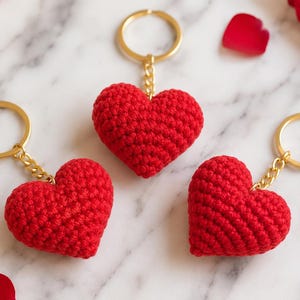 May include: Three red heart-shaped keyrings made of crocheted yarn. Each heart is attached to a gold-coloured metal split ring by a short gold-coloured chain. The texture of the yarn is visible, showing the individual stitches of the crochet work.