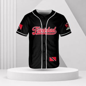 May include: A black baseball jersey with white piping along the button placket and sleeves. The word Trinidad is printed in red script across the chest. The sleeves feature the Trinidad and Tobago flag, and a small emblem is on the left sleeve.