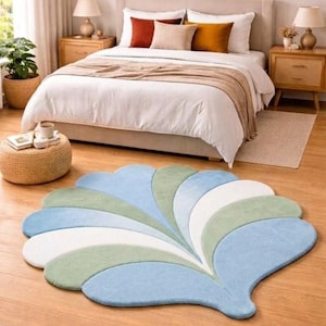 May include: A shell-shaped area rug featuring a fan-like pattern of alternating light blue, sage green, and white segments. The rug is placed on a wooden floor in front of a bed with a white duvet, beige headboard, and two wooden nightstands.