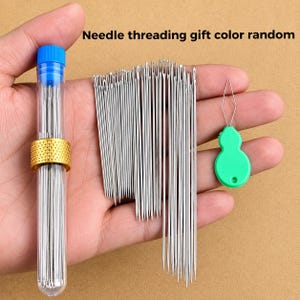 May include: A set of silver sewing needles stored in a clear cylindrical tube with a blue cap and a gold-colored textured band, accompanied by a green plastic needle threader. The text "Needle threading gift color random" is visible.