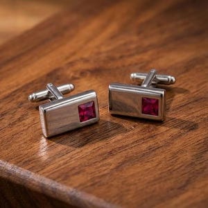 Puede incluir: Un par de gemelos rectangulares de tono plateado, cada uno con una piedra preciosa de color rojo intenso de corte cuadrado incrustada en la superficie de metal pulido. Los gemelos cuentan con un mecanismo de cierre giratorio en la parte posterior.