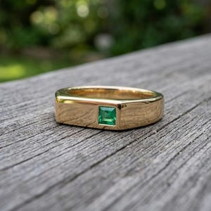 Peut inclure: Une bague en or poli dotée d'une pierre précieuse verte de taille carrée, sertie à fleur au centre de la surface métallique. La bague présente un profil plat et lisse avec un design large qui met en valeur la pierre verte éclatante.