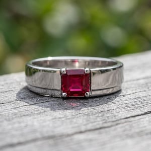 Puede incluir: Un anillo de metal plateado con una piedra preciosa roja vibrante de corte cuadrado engastada en el centro. La banda tiene un acabado liso y pulido con un diseño escalonado que conduce al engaste de cuatro garras que sujeta la piedra.