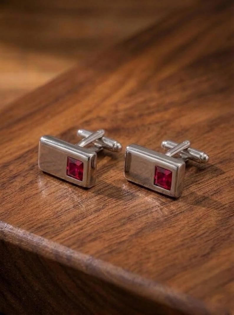 May include: A pair of rectangular silver-toned cufflinks, each featuring a single square-cut deep red gemstone set into one side. The cufflinks have a polished metallic finish and a standard hinged toggle mechanism on the back for secure fastening.