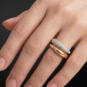 Puede incluir: Un conjunto de dos anillos de oro que se llevan juntos. El anillo superior es una pieza de oro gruesa y curva incrustada con numerosas piedras pequeñas y claras en pavé. El anillo inferior es una pieza de oro lisa y pulida con forma curva.