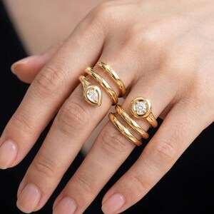 Può includere: Due anelli avvolgenti color oro indossati su dita adiacenti. Un anello presenta una pietra a forma di pera all'estremità di una fascia a spirale. L'altro anello presenta una pietra rotonda taglio brillante incastonata in una lunetta circolare.