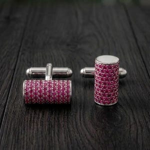 Könnte beinhalten: Ein Paar silberfarbene Manschettenknöpfe mit zylindrischem Design. Die Oberfläche jedes Zylinders ist dicht mit kleinen, runden, leuchtend pinkfarbenen Steinen besetzt, während die Enden und der Befestigungsmechanismus aus poliertem Silbermetall bestehen.
