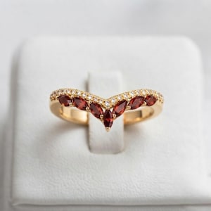 Könnte beinhalten: Ein goldener Ring in Chevron-Form mit einer Reihe tiefroter Granat-Edelsteine im Marquise-Schliff, akzentuiert durch eine Linie kleiner, klarer, runder Steine am oberen Rand. Die Ringschiene ist poliert und folgt einem V-förmigen Design.