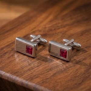 May include: A pair of rectangular silver-toned cufflinks, each featuring a single square-cut deep red gemstone set into one side. The cufflinks have a polished metallic finish and a standard hinged toggle mechanism on the back for secure fastening.