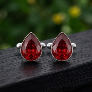 Puede incluir: Un par de gemelos con piedras rojas facetadas en forma de pera engastadas en metal plateado. Cada gemelo presenta un diseño de gota con un soporte metálico pulido y un mecanismo de cierre seguro, mostrando un tono carmesí profundo.