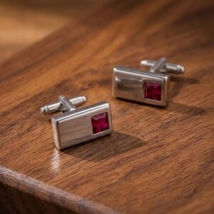 May include: A pair of rectangular silver-toned cufflinks, each featuring a single square-cut deep red gemstone set into the polished metal surface. The cufflinks have a standard hinged toggle mechanism on the back for securing through shirt cuffs.