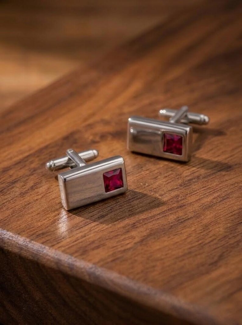 May include: A pair of rectangular silver-toned cufflinks, each featuring a single square-cut deep red gemstone set into the polished metal surface. The cufflinks have a standard hinged toggle mechanism on the back for securing through shirt cuffs.