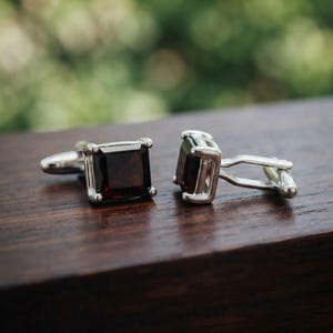 Puede incluir: Un par de gemelos de tono plateado con piedras preciosas de granate rojo oscuro de corte cuadrado. Cada gemelo tiene un marco de metal pulido con un engaste de cuatro garras que sujeta la piedra, unido a un cierre de palanca estándar.