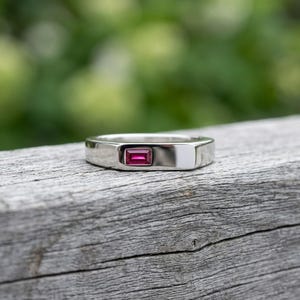 May include: A polished silver-toned metal ring featuring a single rectangular baguette-cut vibrant pink gemstone set horizontally into the centre of the band. The band has a smooth, flat surface and a reflective finish, showcasing a clean design.