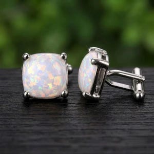 Könnte beinhalten: Ein Paar silberfarbene Manschettenknöpfe mit quadratischen, schillernden weißen Opal-Cabochons. Jeder Manschettenknopf hat einen polierten Metallrahmen mit vier kleinen Krappen, die den Stein halten, und einen klappbaren T-Steg-Verschluss.