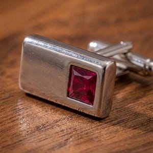 May include: A rectangular silver-toned metal cufflink featuring a smooth, polished surface and a single square-cut, deep red gemstone set into one side. The cufflink includes a standard hinged toggle mechanism on the back for secure fastening.