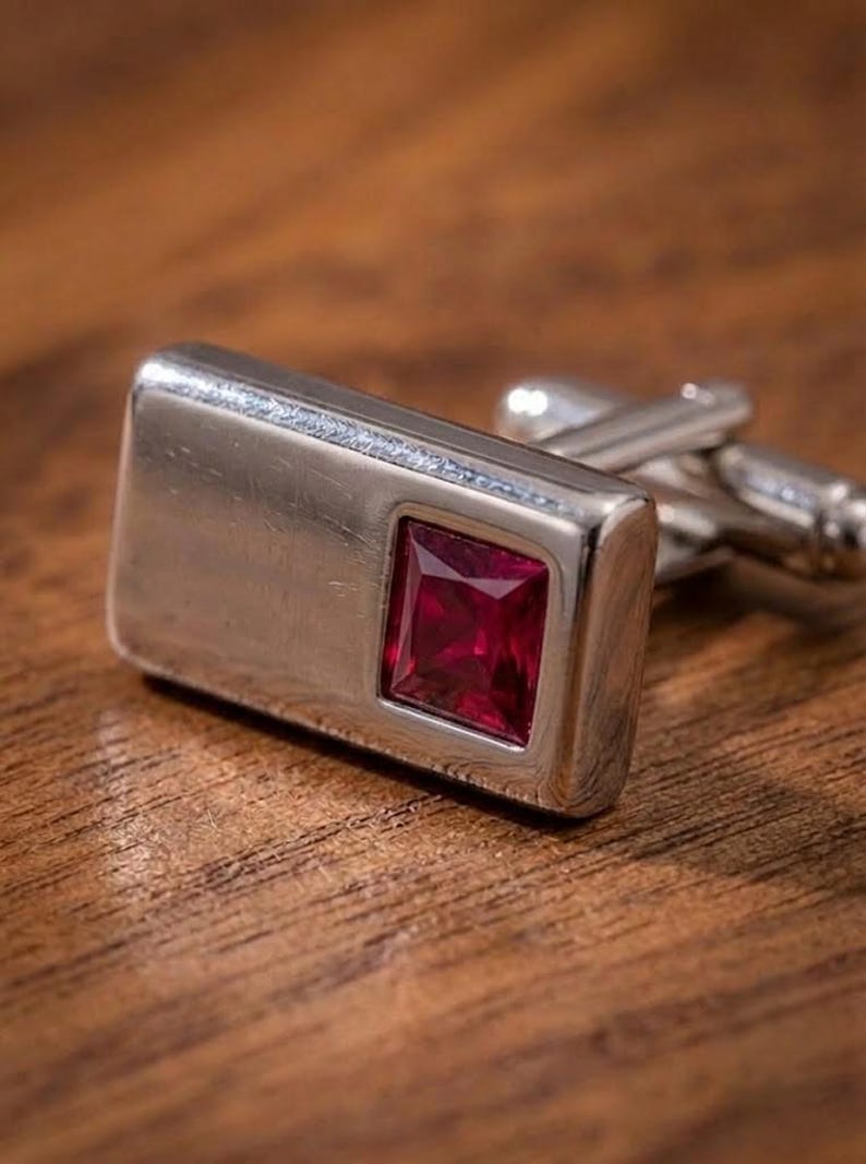 May include: A rectangular silver-toned metal cufflink featuring a smooth, polished surface and a single square-cut, deep red gemstone set into one side. The cufflink includes a standard hinged toggle mechanism on the back for secure fastening.