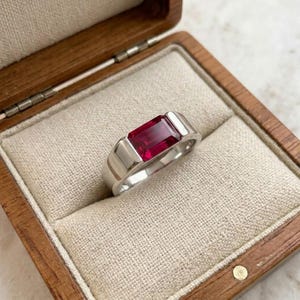 Könnte beinhalten: Ein silberfarbener Ring mit einem rechteckigen, im Smaragdschliff geschliffenen, tiefroten Edelstein, der horizontal in ein glattes, poliertes Metallband gefasst ist. Der Ring liegt in einer mit beigem Stoff ausgekleideten Holzschatulle.