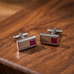 May include: A pair of rectangular silver-toned cufflinks, each featuring a single square-cut, deep red gemstone set into the polished metal surface. The cufflinks have a standard swivel-post toggle mechanism on the back for securing to a shirt cuff.