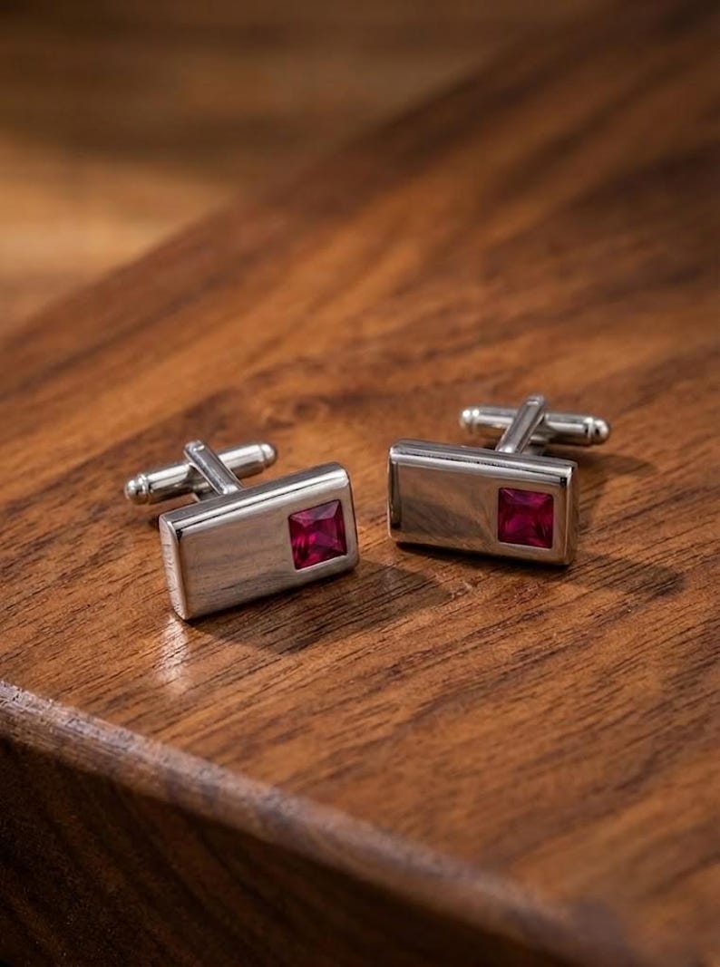 May include: A pair of rectangular silver-toned cufflinks, each featuring a single square-cut, deep red gemstone set into the polished metal surface. The cufflinks have a standard swivel-post toggle mechanism on the back for securing to a shirt cuff.