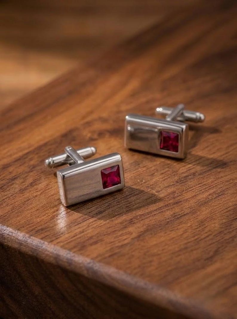 May include: A pair of rectangular silver-toned cufflinks, each featuring a single square-cut deep red gemstone set into the polished metal surface. The cufflinks have a standard hinged toggle mechanism on the back for securing through shirt cuffs.