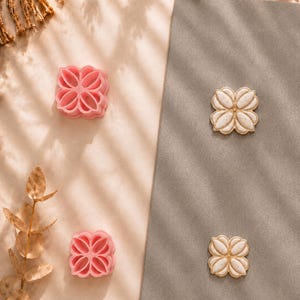 Puede incluir: Dos cortadores de galletas de plástico rosa con forma de flor y dos piezas de arcilla de color blanco roto a juego con bordes dorados. Las piezas de arcilla presentan una textura moteada y un diseño simétrico de cuatro pétalos.