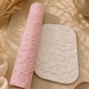 Peut inclure: Un rouleau à pâtisserie cylindrique rose avec un motif floral en relief et un morceau d'argile blanche rectangulaire portant la même empreinte de fleurs et de vignes, illustrant la texture créée par l'outil de gaufrage.