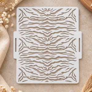 Puede incluir: Una plantilla rectangular blanca con un patrón repetitivo de rayas de tigre. La plantilla tiene una superficie plana con líneas recortadas y dos pequeñas asas rectangulares en los lados izquierdo y derecho, diseñada para manualidades.