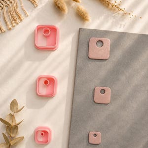 Può includere: Tre taglia-argilla quadrati rosa con inserti centrali circolari disposti verticalmente a sinistra, insieme a tre corrispondenti pezzi di argilla quadrati color rosa polvere con fori circolari su una tavola rettangolare grigia.