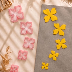 Könnte beinhalten: Fünf rosa blumenförmige Ausstechformen aus Kunststoff in einer vertikalen Reihe sowie fünf gelbe Fondant- oder Teigausstiche in der gleichen Blumenform, die auf einem rechteckigen grauen Brett liegen.