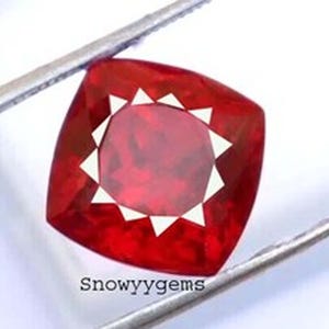 May include: A vibrant red gemstone with a cushion cut, featuring a faceted surface that creates a star-like pattern of light reflections. The stone is held by metal tweezers and has the text Snowyygems printed in black at the bottom.