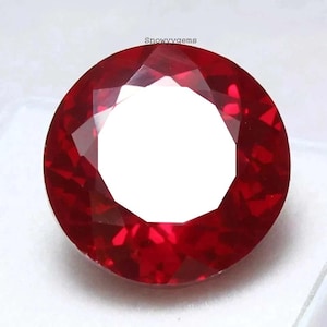 May include: A round, faceted, vibrant red gemstone with a brilliant cut. The stone displays deep crimson hues and sharp geometric facets that reflect light. The text 'Snowyygems' is printed in small black letters above the top edge of the gem.